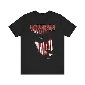 May include: A black short-sleeve t-shirt featuring the word SAMHAIN in red block letters at the top. Below the text is a graphic design showing a face partially obscured by vertical red and white streaks resembling dripping paint or blood.