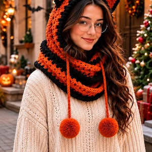May include: A crochet hooded cowl featuring a vibrant orange and black striped pattern with integrated cat-like ears at the top. The accessory includes two long orange braided cords hanging down the front, each finished with a large, fluffy orange pom-pom.