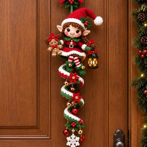 Könnte beinhalten: Ein gehäkelter Weihnachts-Wandschmuck in Form eines Elfen mit einem rot-grünen Spiralschwanz, einem Lebkuchenmann, einer kleinen Laterne und einem weißen Schneeflocken-Ornament. Der Elf trägt eine rote Mütze, ein grünes Kleid und gestreifte Strümpfe.