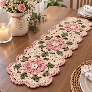 May include: A rectangular crochet table runner featuring a repeating floral pattern with cream-colored yarn, dusty pink petals, and sage green leaves. The runner has a scalloped edge and an intricate openwork design, showcasing detailed needlework.