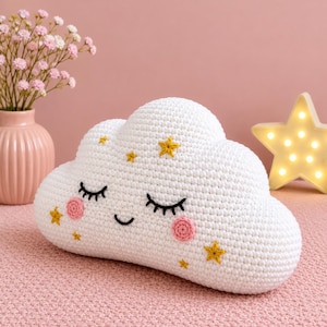 May include: A white crochet cloud pillow featuring a smiling face with closed eyes, embroidered eyelashes, and pink circular cheeks. The cloud is decorated with small yellow embroidered stars and has a soft, textured yarn surface.