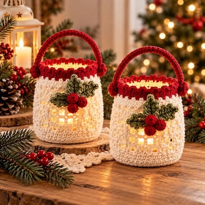 May include: Two cylindrical crochet candle holders featuring a cream-colored body with a red scalloped rim and a red handle. Each holder is decorated with a green crocheted holly leaf and three red berries on the front side.
