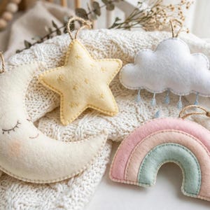 May include: A set of four felt hanging ornaments featuring a cream crescent moon with a stitched face, a pale yellow star with gold embroidery, a white cloud with blue raindrops, and a pastel rainbow in shades of pink, peach, and mint green.