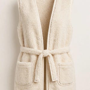 May include: A sleeveless cream-colored vest made of textured fleece material. The vest features an open front design with a matching tie belt at the waist and two patch pockets on the front. The edges are finished with smooth trim.