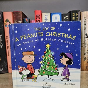 May include: A blue book titled "The Joy of A Peanuts Christmas" featuring Charlie Brown, Snoopy, Lucy, and a decorated Christmas tree. The cover has white stars and the text "50 Years of Holiday Comics!" Hallmark Books.
