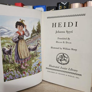 May include: An open book with the title "HEIDI" by Johanna Spyri. The illustration shows a girl in traditional clothing with goats in a field. The book is part of the Illustrated Junior Library series.