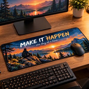 May include: A rectangular desk mat featuring a landscape illustration of a person sitting on a rock overlooking a lake at sunset, with the text "MAKE IT HAPPEN - one day at a time" printed in white and yellow across the top center of the mat.