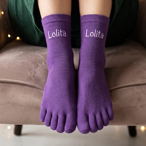 May include: A pair of purple toe socks featuring the word Lolita embroidered in white thread on the front of each sock. The socks are designed with individual compartments for each toe and reach mid-calf, covering the feet completely.