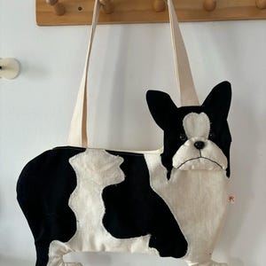 May include: A black and white tote bag shaped like a French Bulldog. The bag features a cream-colored canvas strap, a plush-style dog head with black ears, and a body with irregular black and white patches. A small red tag is visible on the side.