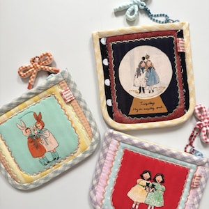 May include: Three square fabric pouches with zipper closures and gingham borders. Each pouch features a unique illustration of two figures, decorative stitching, a small 'HAND MADE' tag, and a matching fabric bow attached to a ball chain.