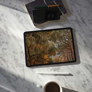May include: A tablet displaying a forest scene with the text The Open Path Notebook, a black smartphone, a green and gold pen, a marble-patterned notebook, a white stylus, and a white ceramic mug filled with dark coffee on a white marble surface.