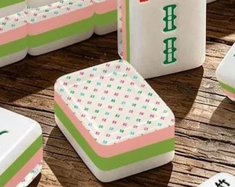 Könnte beinhalten: Ein rechteckiger Mahjong-Spielstein mit einer weißen Oberseite, die ein wiederkehrendes Muster aus kleinen grünen und rosa Kreuzen aufweist. Die Seiten des Spielsteins sind mit geschichteten weißen, grünen und rosa Bändern versehen.