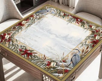 Könnte beinhalten: Ein quadratischer Holztisch mit einer Tischplatte, die eine Winterlandschaft zeigt. Der Rand ist mit einem festlichen Muster aus grünen Tannenzweigen, roten Beeren, Tannenzapfen und roten Schleifen sowie einem blauen Reiher verziert.