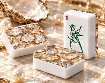 Könnte beinhalten: Drei rechteckige Mahjong-Spielsteine mit weißer Basis und Oberseiten aus eingelassenen, schillernden Muschelfragmenten mit goldfarbenen Rändern. Ein Stein steht aufrecht und zeigt eine rote Eins und ein grünes Vogelmotiv.