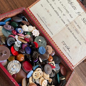 May include: A rectangular box filled with a large collection of assorted buttons in various colours, shapes, and sizes. The inside lid features a vintage certificate dated 20 SEP 1934, certifying that Derek Green, aged 11, has shown ability to swim 25 yards breast stroke.