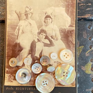 May include: A collection of assorted round buttons in varying sizes and shades of white, cream, and iridescent grey, arranged over a vintage sepia-toned photograph of three people, with the text "From NIGHTINGALE American Fine Art Photo Studio COOKTOWN".
