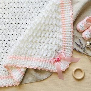 May include: A white crocheted blanket featuring a textured stitch pattern, a light pink border, and a matching pink satin ribbon bow. Beside the blanket are a pair of white crocheted baby booties, a small silver rattle, and a wooden ring.