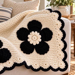 May include: A cream-colored crochet blanket featuring large black flower motifs with central cream-colored petals. The blanket has a decorative scalloped border in alternating black and cream yarn, creating a textured, layered appearance.