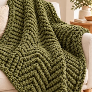 May include: A chunky knit throw blanket in a deep olive green color featuring a prominent chevron pattern. The blanket is constructed from thick, soft yarn loops, creating a textured surface that drapes over the arm of a light-colored sofa.