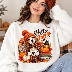 Könnte beinhalten: Ein weißes Sweatshirt mit einer Grafik eines Beagles, der eine orangefarbene Strickmütze und einen karierten Schal trägt und eine Tasse mit Pfotenabdruck hält. Das Design zeigt Kürbisse, Herbstblätter, eine Laterne und Bücher mit Aufschriften.