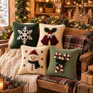May include: Four square crochet throw pillows featuring holiday designs. One dark green with a white snowflake, one cream with red bells, one cream with a snowman, and one dark green with two candy canes. Each pillow has a textured knit surface.