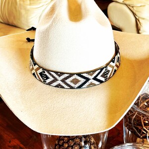 May include: A cream-colored cowboy hat with a decorative beaded band featuring a geometric pattern in black, white, and gold. The hat is set against a warm, neutral background, showcasing its classic Western style.