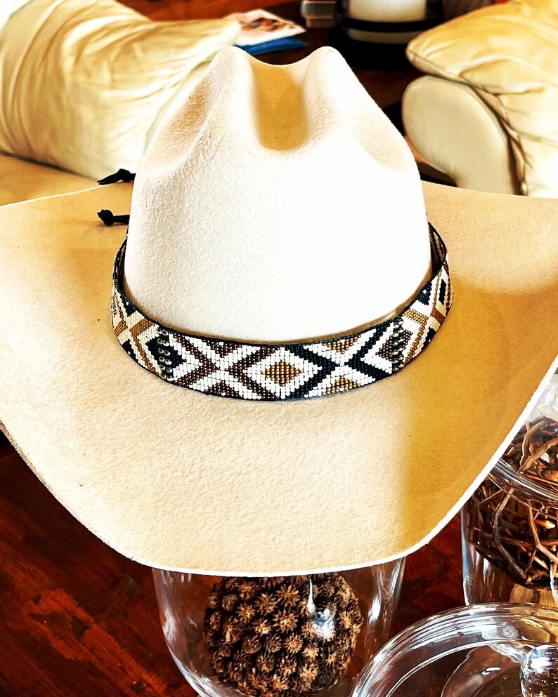 May include: A cream-colored cowboy hat with a decorative beaded band featuring a geometric pattern in black, white, and gold. The hat is set against a warm, neutral background, showcasing its classic Western style.
