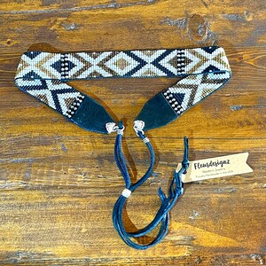 May include: A beaded belt with a geometric pattern in white, black, and gold. The belt has dark blue leather ends and matching blue leather ties. The tag reads "Fleurdesignz Western Jewelry Proudly Handmade in the USA."
