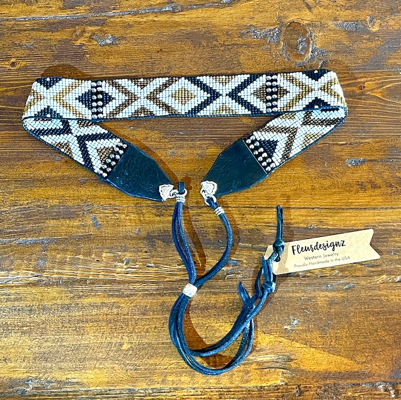 May include: A beaded belt with a geometric pattern in white, black, and gold. The belt has dark blue leather ends and matching blue leather ties. The tag reads "Fleurdesignz Western Jewelry Proudly Handmade in the USA."