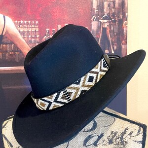 May include: A black cowboy hat with a decorative beaded band featuring a geometric pattern in white, black, and gold. The hat is displayed on a mannequin head. The hat's brim is curved.