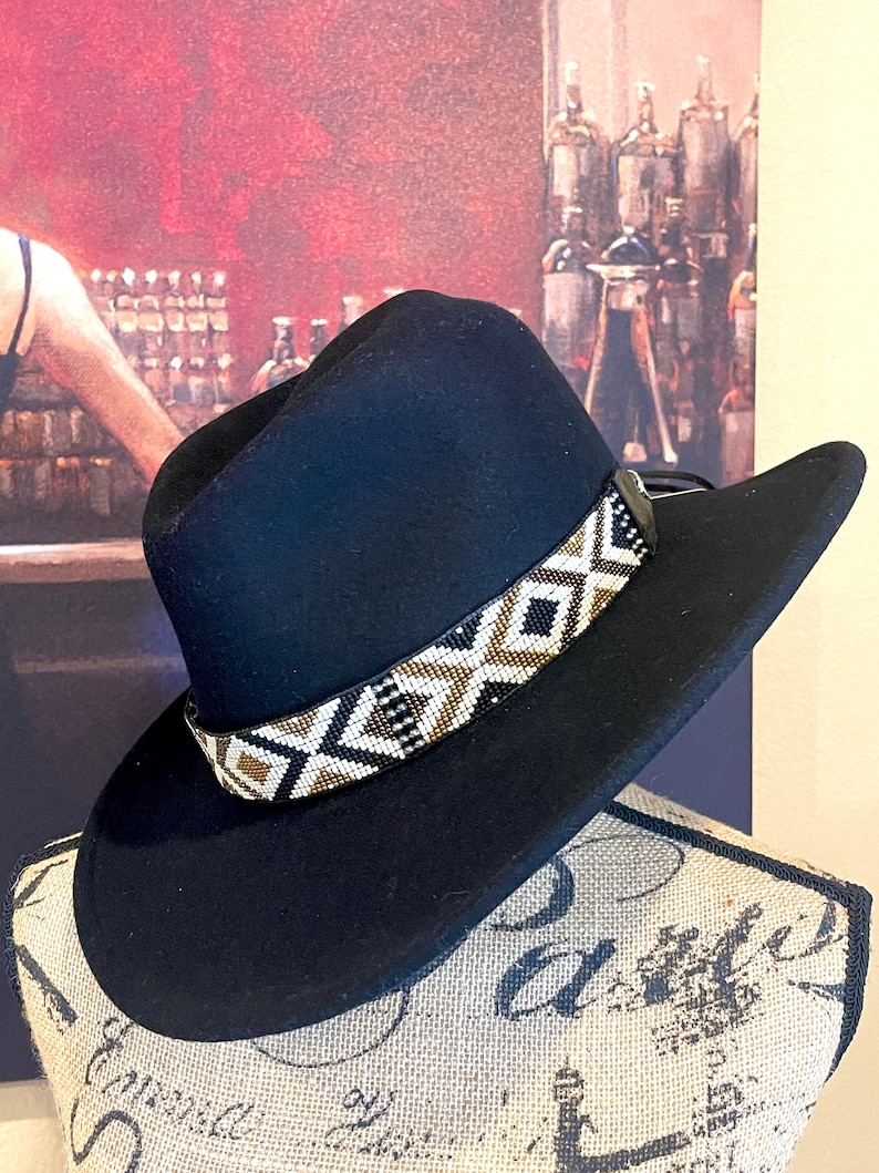 May include: A black cowboy hat with a decorative beaded band featuring a geometric pattern in white, black, and gold. The hat is displayed on a mannequin head. The hat's brim is curved.