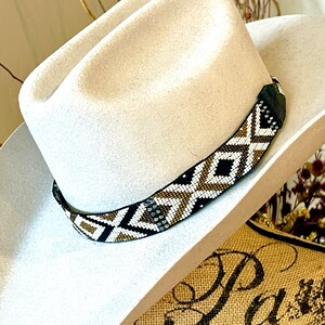 May include: A cream-colored cowboy hat with a beaded hatband. The hatband features a geometric pattern in black, white, and gold. The hat is made of a soft material and has a classic western style.