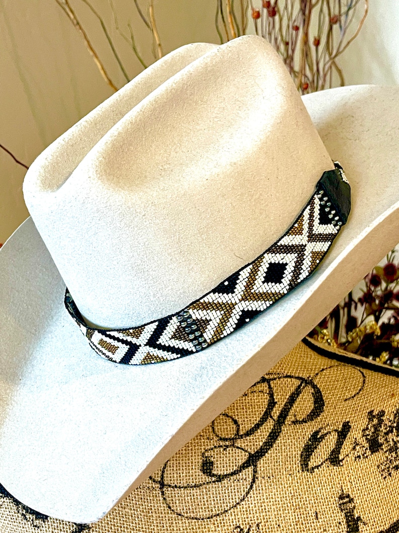May include: A cream-colored cowboy hat with a beaded hatband. The hatband features a geometric pattern in black, white, and gold. The hat is made of a soft material and has a classic western style.
