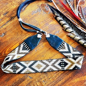 May include: A beaded headband with a geometric pattern in black, white, and gold. The headband has a dark blue leather strap and is adorned with small silver-colored studs. Feathers in various colors are visible in the background.