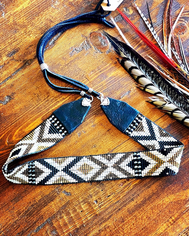 May include: A beaded headband with a geometric pattern in black, white, and gold. The headband has a dark blue leather strap and is adorned with small silver-colored studs. Feathers in various colors are visible in the background.