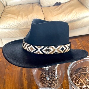 May include: A black fedora-style hat with a wide brim. The hat features a decorative band with a geometric pattern in white, brown, and gold beads. The hat is displayed on a glass surface.