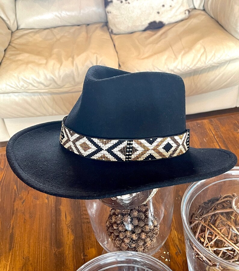 May include: A black fedora-style hat with a wide brim. The hat features a decorative band with a geometric pattern in white, brown, and gold beads. The hat is displayed on a glass surface.