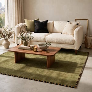 Peut inclure: Un canapé texturé couleur crème avec trois coussins décoratifs olive, noir et crème. Une table basse rectangulaire en bois repose sur un grand tapis vert olive avec une bordure à motifs noirs et crème, supportant un bol et des livres.