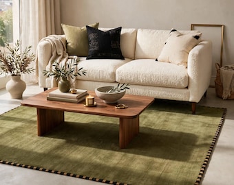 Peut inclure: Un canapé texturé couleur crème avec trois coussins décoratifs olive, noir et crème. Une table basse rectangulaire en bois repose sur un grand tapis vert olive avec une bordure à motifs noirs et crème, supportant un bol et des livres.