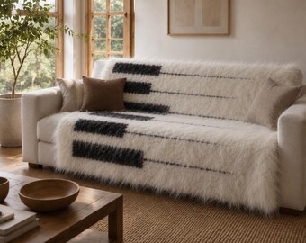 Peut inclure: Un jeté de canapé en fausse fourrure blanche orné d'un motif de touches de piano noires et blanches. La texture à poils longs et hirsutes recouvre l'assise, accompagnée de deux coussins unis dans des tons marron clair et beige.