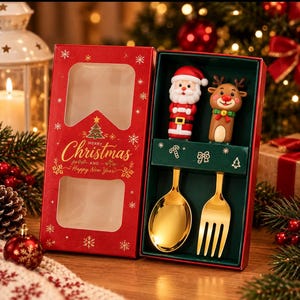 Puede incluir: Un juego de cuchara y tenedor dorados dentro de una caja de regalo roja. El mango de la cuchara tiene una figura de Santa Claus y el del tenedor una de reno. La tapa de la caja tiene una ventana y el texto Merry Christmas and Happy New Year.