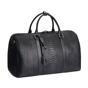 Könnte beinhalten: Eine schwarze Reisetasche mit einer strukturierten Schlangenhaut-Optik auf dem Hauptteil, ergänzt durch glatte schwarze Lederbesätze, zwei Tragegriffe, einen Reißverschluss oben und einen kleinen passenden Kofferanhänger am Griff.