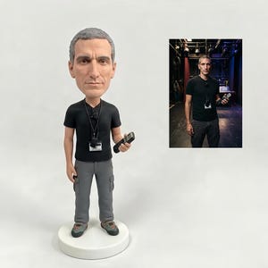 May include: A custom bobblehead figurine depicting a person with short gray hair, wearing a black t-shirt, gray cargo pants, and a lanyard with a badge. The figure holds a black remote control in one hand and stands on a round white base.