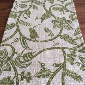 May include: A rectangular table runner with a botanical design. The runner features a light beige background with a repeating pattern of green vines, leaves, and floral elements. The fabric has a textured appearance, adding visual interest to the design.