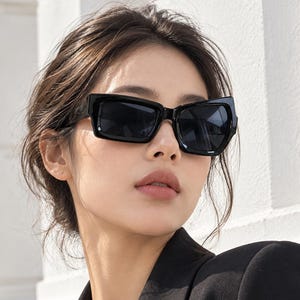 Puede incluir: Gafas de sol rectangulares negras con monturas gruesas y lentes oscuras. El diseño presenta una forma angular audaz con patillas anchas y un acabado liso y brillante, proporcionando una cobertura completa para los ojos en una silueta.