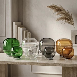 May include: A collection of eight glass vases arranged in pairs of varying sizes and colors, including green, clear, grey, and amber. Each vase features a rounded, bulbous body with a short, cylindrical base and a wide, open top opening.