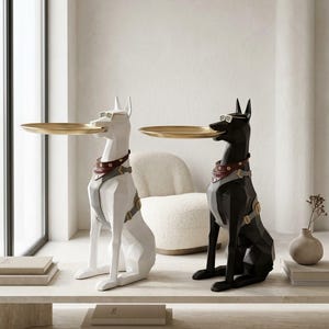 May include: Two geometric dog sculptures, one white and one black, each wearing sunglasses and a red and gray harness. Each dog holds a round, gold-colored metal tray in its mouth. The figures are seated in an upright, alert posture.