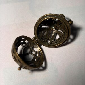 May include: An open, spherical locket made of antique brass-toned metal. The two halves are connected by a small hinge and feature a detailed, intricate cut-out filigree pattern throughout the surface, revealing the hollow interior space.