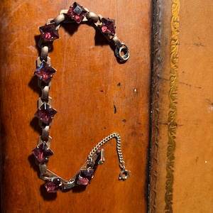 May include: A metal link bracelet featuring seven square-cut, deep purple faceted stones set in individual metal frames. The bracelet includes a short, fine-link safety chain attached to a circular spring ring clasp at one end of the piece.
