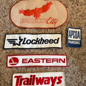 May include: A collection of five embroidered patches featuring various logos. The top patch is oval-shaped with a red eagle and the text BUICK City. Below are rectangular patches with the text Lockheed, APCOA PARKING, EASTERN, and Trailways.